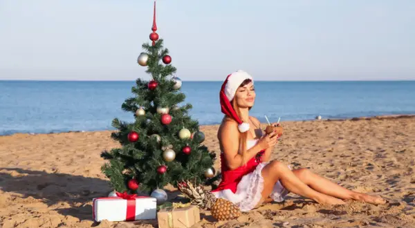 Kobieta w stroju Mikołaja pije koktajl na plaży obok choinki. Czy 28 grudnia jest dniem wolnym od pracy?