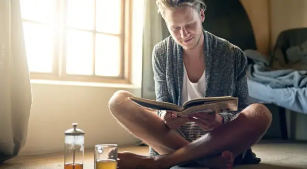 Hombre joven leyendo un libro, disfrutando del **slow living** junto a una ventana con luz natural y una taza de té.