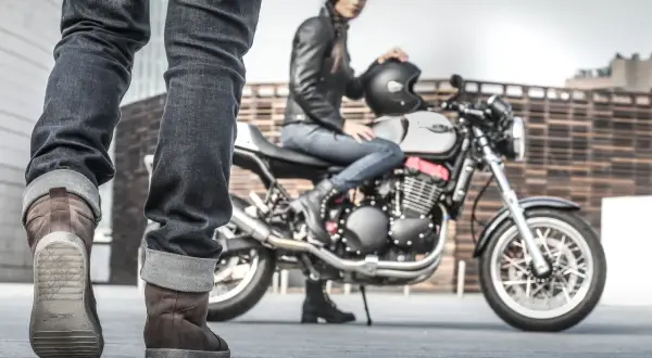 Kobieta w skórzanej kurtce siedzi na motocyklu, obok stoją nogi w brązowych butach motocyklowych.