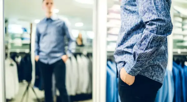 Mann im Spiegel prüft seinen lässigen kleidungsstil herren. Blaues Hemd mit Muster, schwarze Hose.