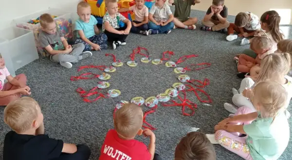 Dzieci siedzą w kółku na dywanie, patrząc na okrąg z zawieszkami. Może to być część zabawy, jak w "Mam chusteczkę haftowaną".