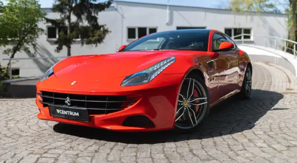 Czerwone Ferrari FF na brukowanej drodze, z budynkiem i drzewami w tle.