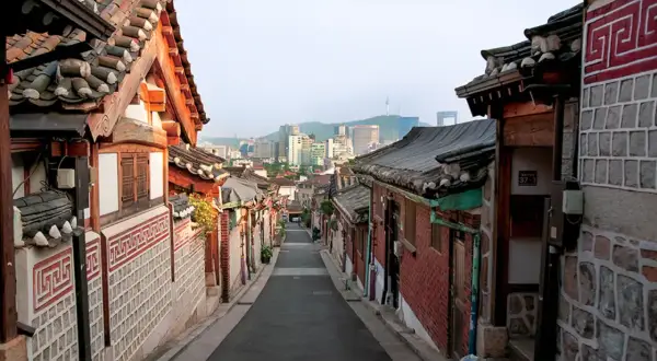 En smal gata kantad av traditionella hus med tegeltak leder mot en modern stadssilhuett i Seoul, Sydkorea.