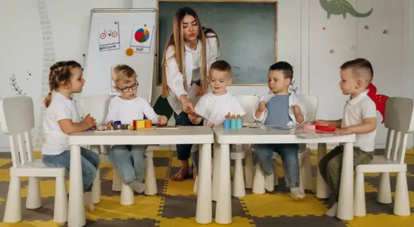 Nauczycielka bez wykształcenia pedagogicznego prowadzi zajęcia z dziećmi w przedszkolu. Dzieci siedzą przy stolikach, bawiąc się klockami i kartami.