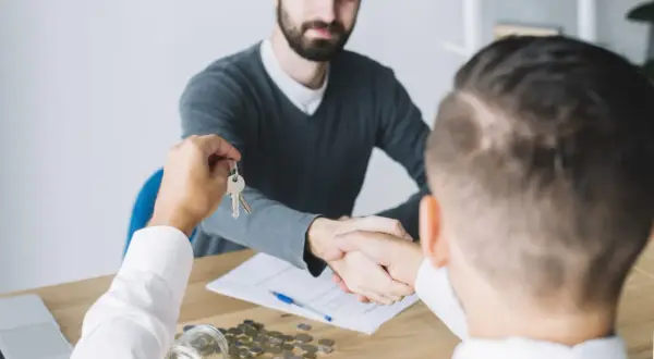 Conclusione di una trattativa acquisto casa con agenzia: scambio chiavi e stretta di mano. Sul tavolo, monete e documenti.