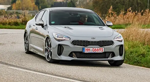 Szary Kia Stinger z kierowcą za kierownicą jedzie po krętej drodze, otoczony jesienną roślinnością.