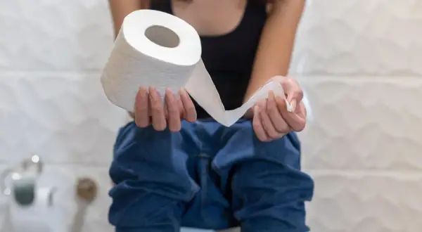 Osoba siedząca na toalecie trzymająca rolkę papieru toaletowego, sugerująca problemy z biegunką.