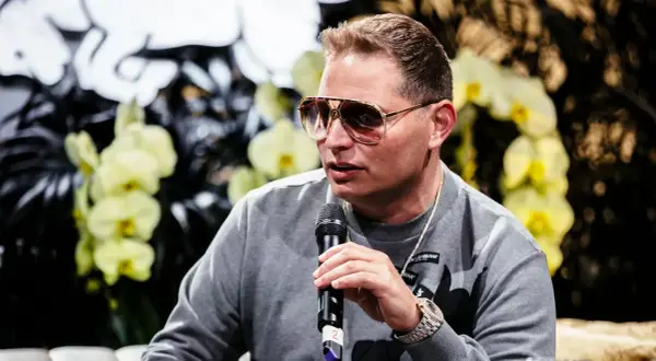 Scott Storch, known for his hit songs, speaks into a microphone, wearing sunglasses and a grey sweatshirt.