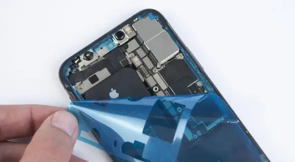 Dłoń odkleja niebieską folię od wnętrza iPhone'a XR z widoczną baterią.