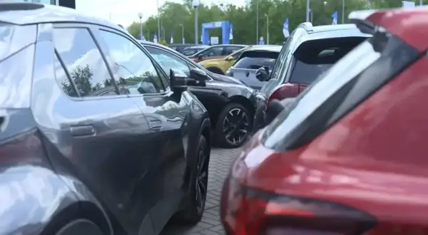 Rzędy samochodów, w tym czerwony i szary SUV, czekają na swoich właścicieli. W tle widać bramę z logo, sugerującą salon samochodowy lub wydarzenie motoryzacyjne.
