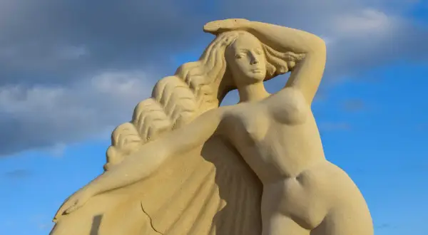 Zandstenen beeld van een **Griekse godin** met golvend haar, tegen een blauwe lucht met wolken.