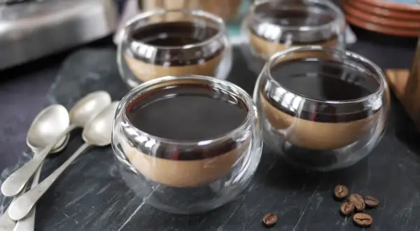 Fyra små glas med kaffe pannacotta, toppad med mörk sirap. Tre skedar ligger till vänster, och kaffebönor sprider sig på stenen.