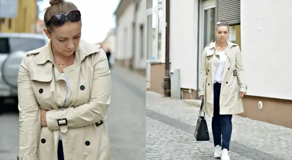 Kobieta w beżowym trenczu i sportowych stylizacjach, z torebką i okularami przeciwsłonecznymi, idzie po brukowanej ulicy.