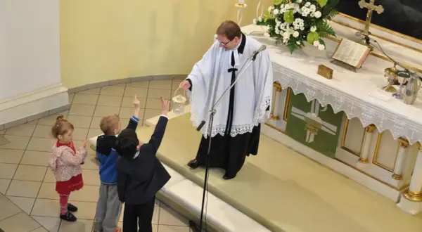 Dzieci z podniesionymi rękami słuchają księdza. Różnice między ewangelikami a katolikami są widoczne w liturgii i symbolach.