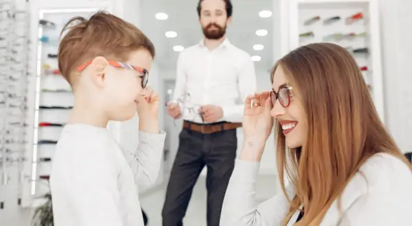 Niños probándose gafas en una óptica. La madre ayuda a su hijo con un **ojo vago** a elegir montura.