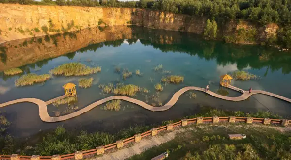 Malowniczy park Gródek z drewnianą kładką wijącą się pośród turkusowej wody i trzcin. W oddali skalne zbocza i las.