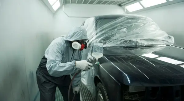 Un operaio in tuta protettiva e maschera spruzza vernice su un'auto scura, parzialmente coperta da un telo trasparente. L'auto sembra rovinata, in fase di restauro.