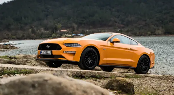 Pomarańczowy Mustang 2.3 Ecoboost z czarnymi felgami stoi nad jeziorem, gotowy na przygodę.