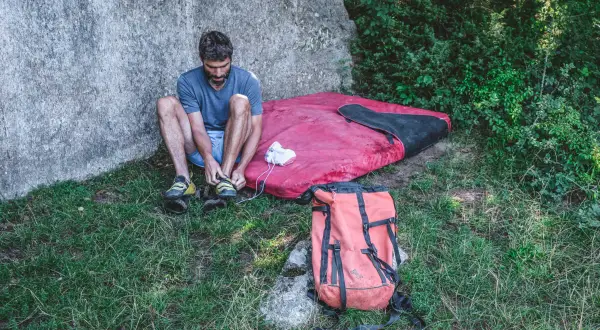 Man förbereder sig för kjugekull bouldering, spänner sina skor bredvid en tjock madrass och en orange ryggsäck.