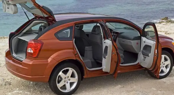 Orange Dodge Caliber 2007 med öppna dörrar och baklucka vid havet. Perfekt för en roadtrip!