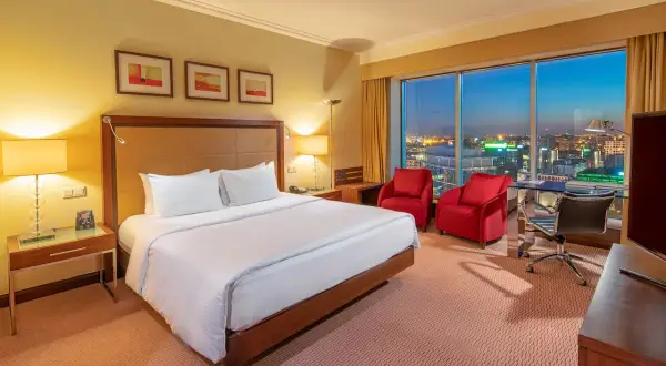 Przytulny pokój w hotelu Hilton Warszawa z widokiem na nocne miasto. Duże łóżko, dwa czerwone fotele i biurko.