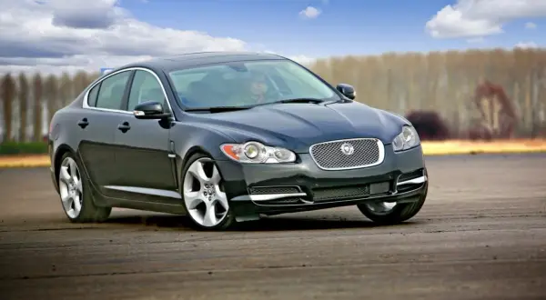 Czarny Jaguar XF na torze. Kierowca w środku, chmury na niebie. Wiele pozytywnych jaguar xf opinie potwierdza jego sportowy charakter.