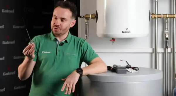 Mężczyzna w zielonej koszulce prezentuje element, który może być anodą tytanową, obok zasobnika wody.