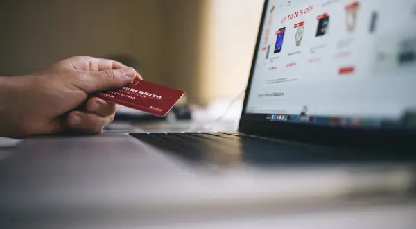 Dłoń trzyma czerwoną kartę przy laptopie z otwartą stroną zakupową. Czy debet jest oprocentowany? To pytanie zadaje sobie wiele osób podczas zakupów online.