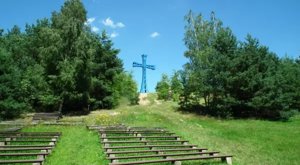 Niebieski krzyż na wzgórzu, najwyższy szczyt wielkopolski, otoczony drzewami. Poniżej amfiteatr z drewnianymi ławami.