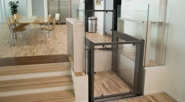 Piattaforma elevatrice per interni con cabina in vetro e struttura metallica, integrata in un ambiente moderno con scale in legno e sala da pranzo.