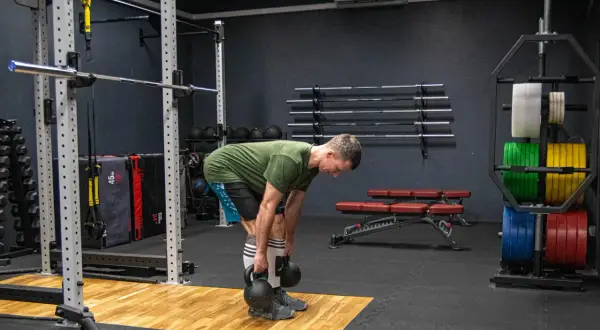 Mężczyzna wykonuje martwy ciąg rumuński z kettlebellami, ćwicząc mięśnie tylnej taśmy. W tle widać sprzęt do treningu siłowego, w tym sztangi i obciążenia.