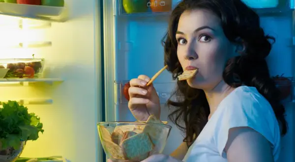 Une femme, prise par une **sensation de faim**, grignote des biscuits devant un réfrigérateur ouvert, rempli de fruits et légumes.