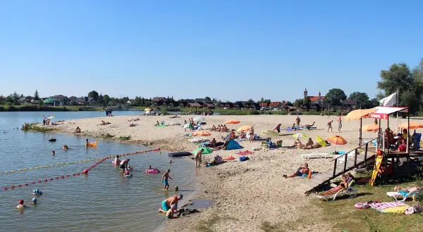 Słoneczny dzień na piaszczystej plaży bobrowego rozlewiska w Zabierzowie Bocheńskim. Ludzie opalają się, kąpią i bawią w wodzie.