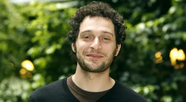 Claudio Santamaria giovane, con capelli ricci e barba, sorride con uno sfondo verde sfocato.