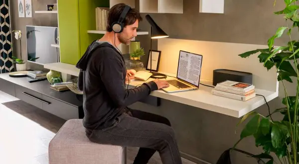 Uomo con cuffie lavora al laptop in un angolo studio in soggiorno. Libri, lampada e pianta completano l'arredo.