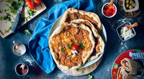 Lahmacun rezept original: Fladenbrot mit Hackfleisch, Tomaten, Petersilie und Käse, daneben Oliven und ein Wrap.