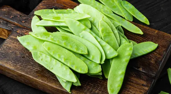 Un mucchio di taccole fresche, verdi e piatte, su un tagliere di legno scuro. Cosa sono? Deliziose!