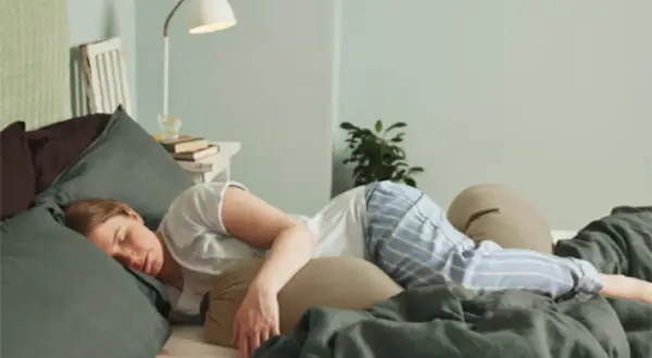 Kvinna sover i sängen, omgiven av kuddar. Hon har på sig randiga pyjamasbyxor. Kanske hon känner av slempropp eller flytning.