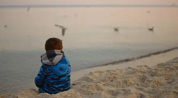 Chłopiec w niebieskiej bluzie siedzi na plaży, obserwując mewy. Spokojna atmosfera nad morzem może pomóc w tym, jak wyciszyć dziecko nadpobudliwe.