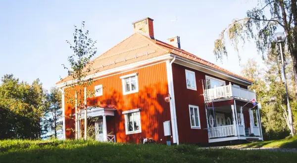 Rött hus med vita knutar och veranda, omgivet av grönska. Ett charmigt vandrarhem i Jokkmokk som bjuder in till en naturnära vistelse.