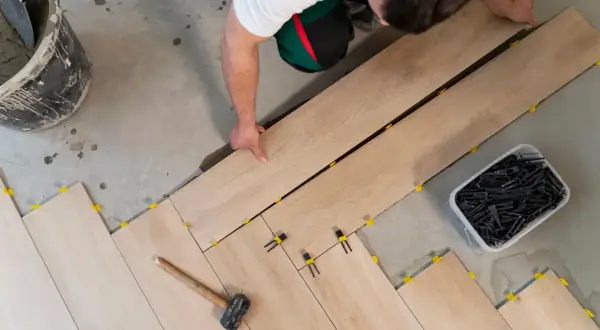 Pose d'un carrelage imitation bois sur un sol en béton. Un ouvrier ajuste les lames, des cales jaunes et noires sont visibles.