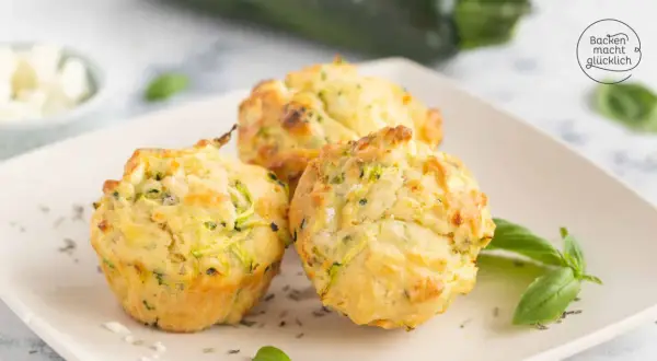 Drei goldbraune Zucchini-Muffins mit Kräutern und Käse auf einem Teller, garniert mit Basilikum.