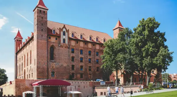 Imponujące ceglane zamki do zwiedzania z czerwonymi dachami i wieżami. Wokół zielony trawnik i drzewa.