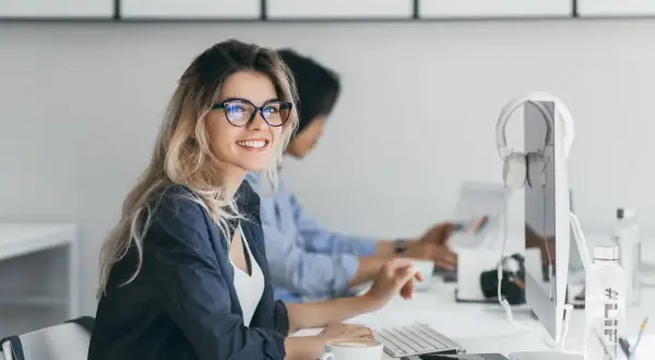 Uśmiechnięta kobieta w okularach pracuje przy komputerze. W tle koleżanka. To obrazek idealnie oddający ducha dyrektywy work life balance.