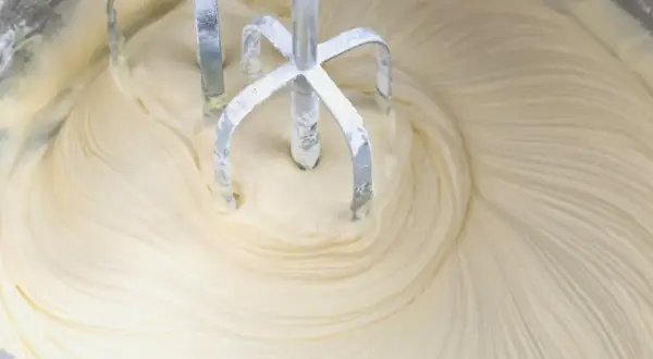 Mieszadła miksera obracają się w gładkim, kremowym **kremie z mascarpone i białej czekolady**, tworząc wiry.