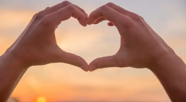 Hände formen ein Herz vor einem Sonnenuntergang. Eine spirituelle Person drückt Liebe und Verbundenheit aus.