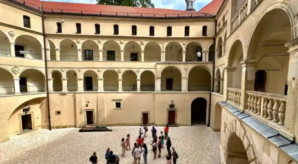 Dziedziniec zamku z arkadowymi krużgankami, kamienna posadzka przypomina pieskowa skała. Grupa turystów zwiedza historyczne miejsce.