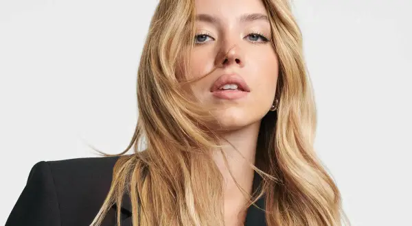 Sydney Sweeney, con su cabello rubio ondulado y un blazer negro, mira directamente a la cámara con una expresión cautivadora.