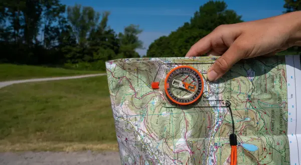 Dłoń trzyma mapę i kompas, wskazując kierunki północ południe wschód zachód. W tle zielony las i błękitne niebo.