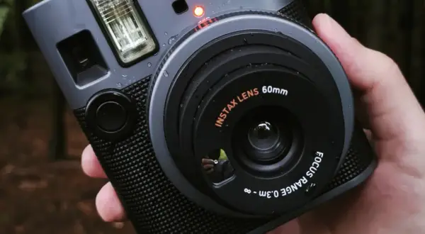 Szary aparat natychmiastowy Instax z obiektywem 60mm, trzymany w dłoni na tle lasu.